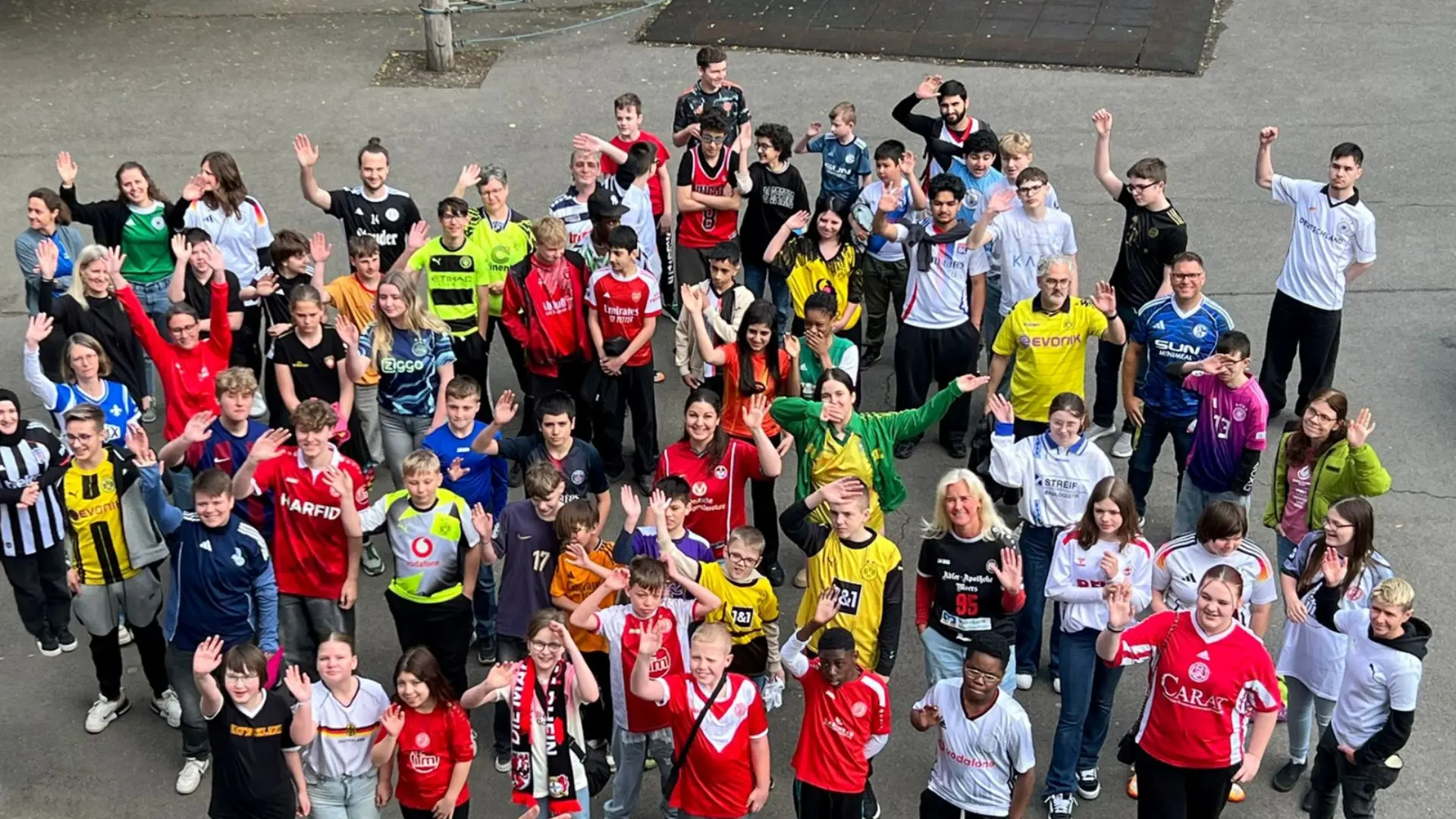 Eine große Gruppe aus Schüler*innen und Lehrkräften, die alle verschiedene Trikots tragen, in die Kamera schauen und winken