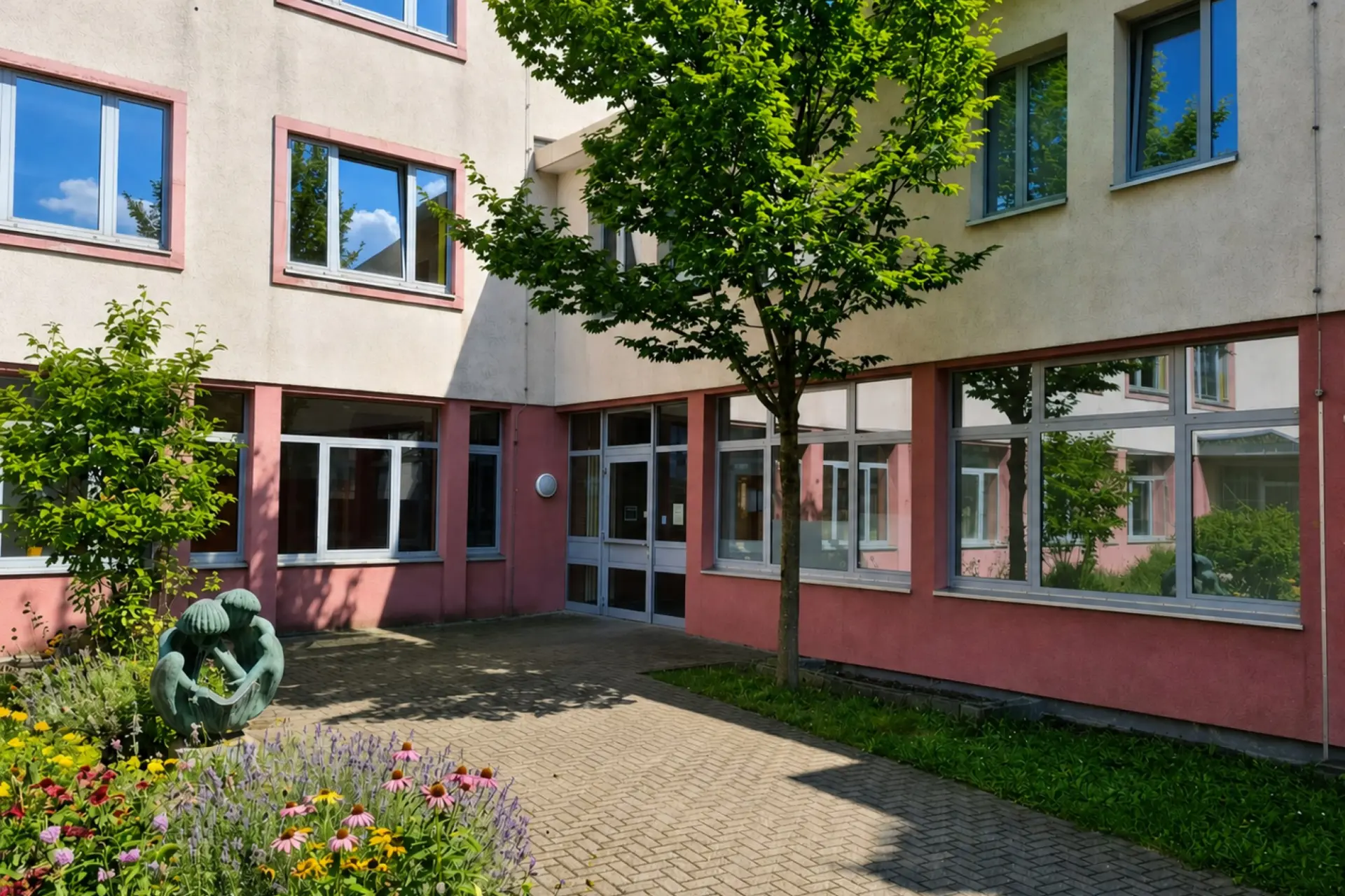 Gepflasterter Vorplatz und Haupteingang der LVR-Wilhelm-Körber-Schule mit einer Skulptur im Vordergrund und hohen Fensterfronten im rosa-beigen Schulgebäude