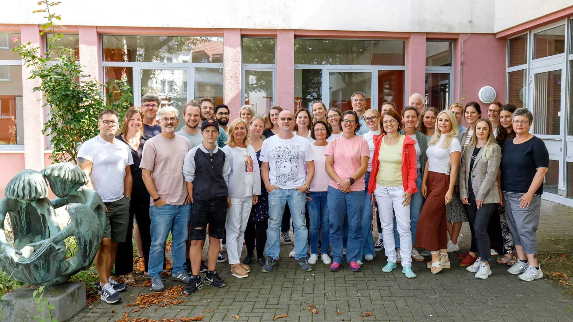 Ein Gruppenporträt von mehreren Personen, die das Schulkollegium der LVR-Wilhelm-Körber-Schule bilden. Sie wurden an einem sonnigen Tag vor dem Schulgebäude fotografiert.