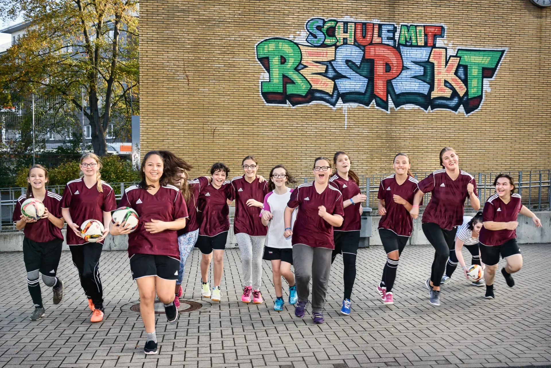 Eine Gruppe aus 13 Mädchen mit Fußballtrikots und manche mit einem Fußballball in der Hand rennen lachend auf die Fotokamera zu.