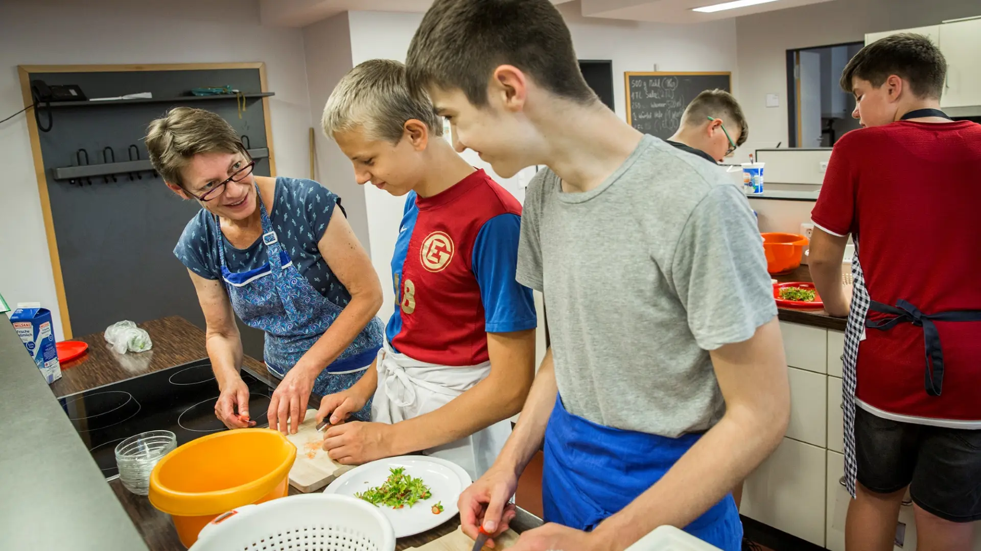 Zwei Schüler schneiden Erdbeeren an einer Theke in der Schulküche. Neben ihnen ist eine Lehrkraft, die etwas zu ihnen sagt. Hinter ihnen ist noch eine Küchenzeile zu sehen, da arbeiten zwei weitere Schüler.