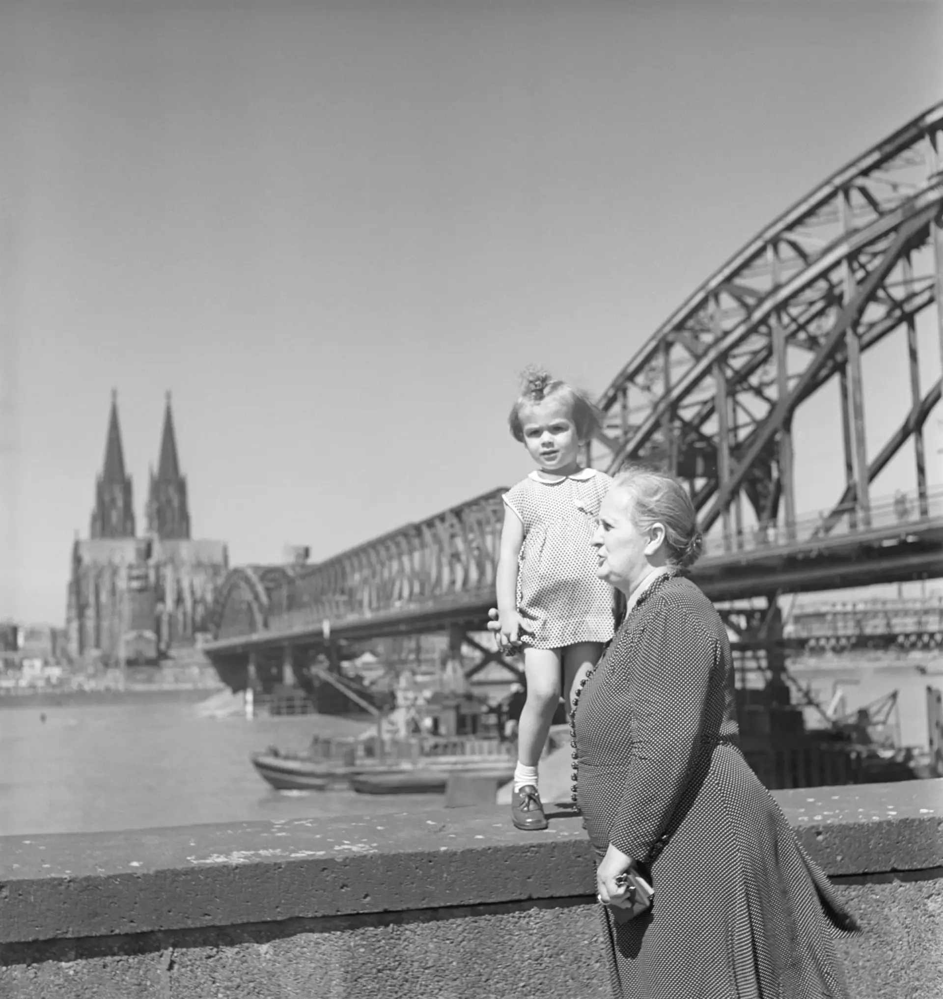 Ein Mädchen steht auf einer Mauer; im Hintergrund sieht man einen Fluss, eine Brücke und eine Kirche.