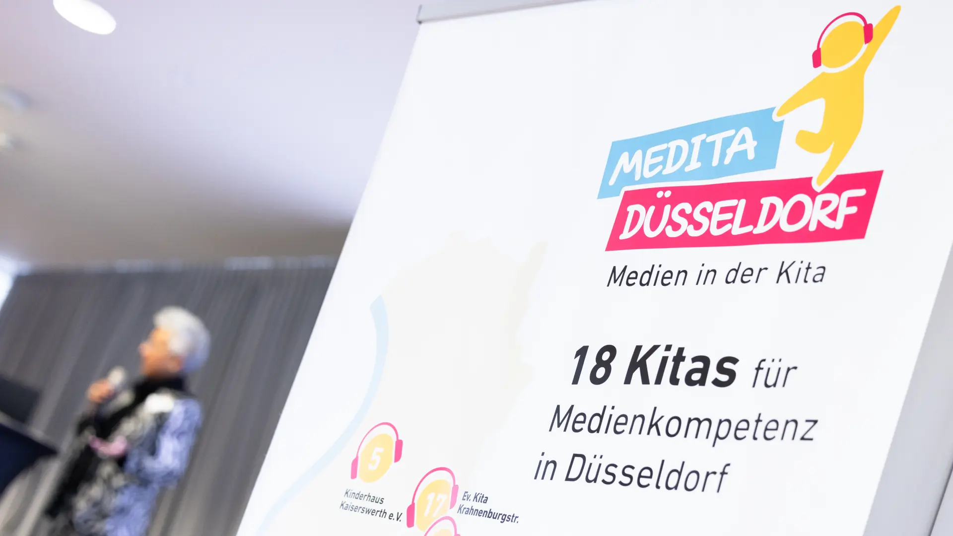MEDITA-RollUp im Vordergrund, Moderatorin auf Bühne im Hintergrund