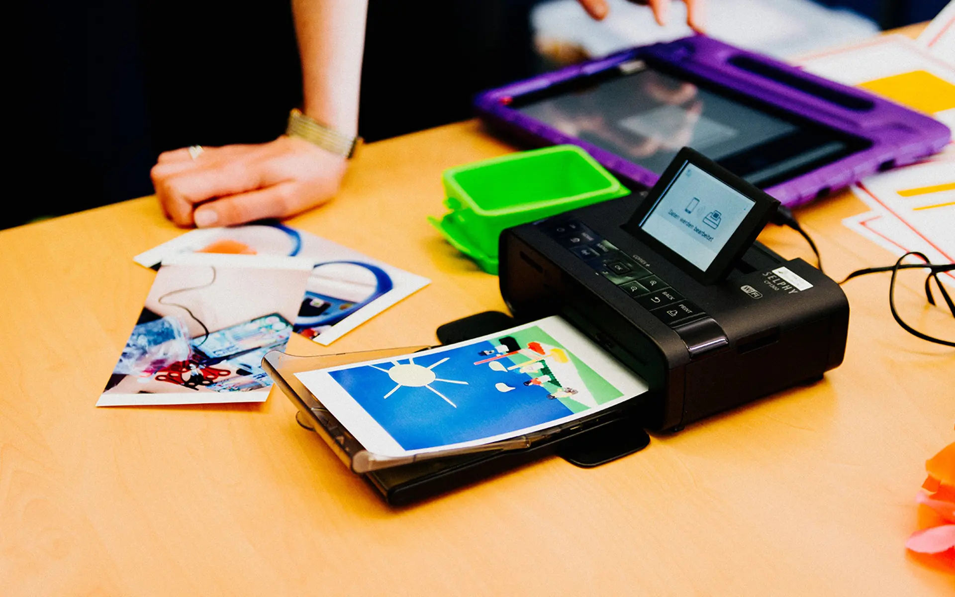 Im Vordergrund steht ein kleiner Fotodrucker, im Hintergrund liegen ausgedruckte Fotos, ein Ipad und andere Materialien