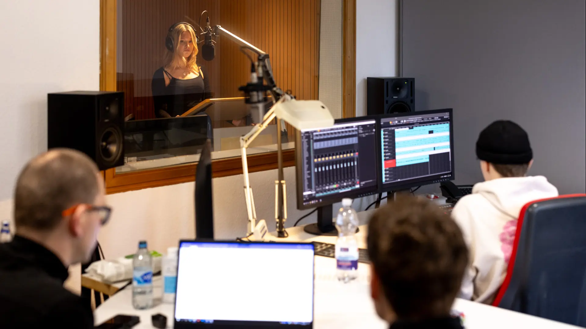 Drei Personen sitzen vor Audio- und Schnittmonitoren in einem Studio, während hinter einer Glasscheibe eine Sprecherin mit Kopfhörern in ein Mikrofon spricht.