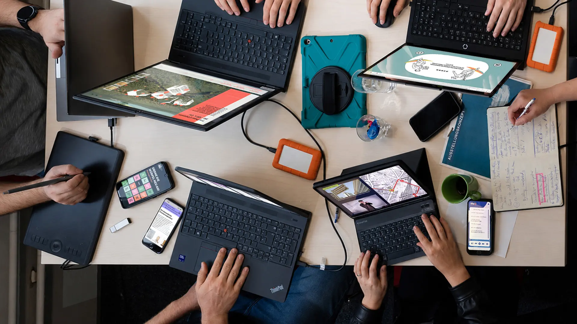  An einem tisch arbeiten mehrere Personen an Medienprojekten, man sieht nur ihre Hände und mehrere Laptops und Handys