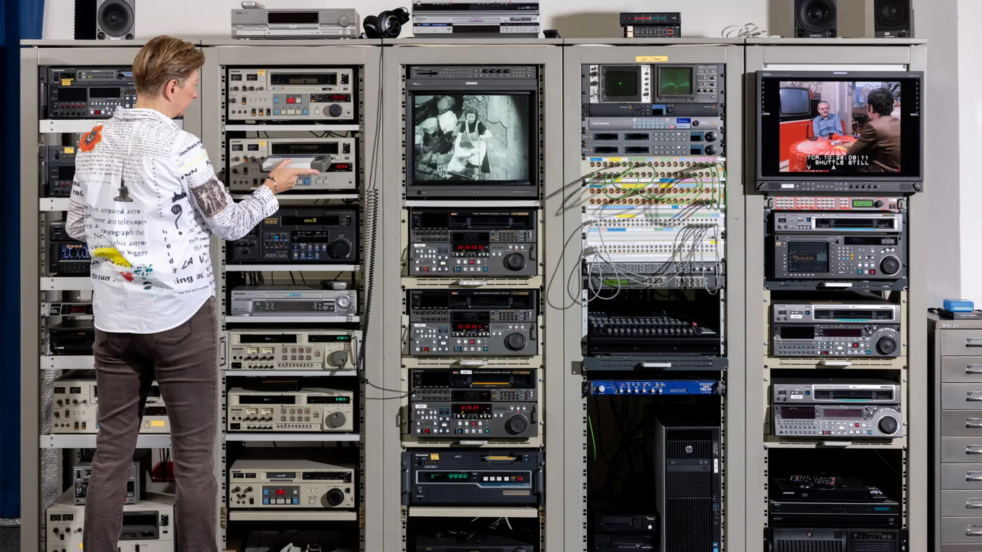 Eine Person bedient in einem Technikraum mehrere große Regale mit Video- und Audiogeräten, während auf Monitoren Filmszenen zu sehen sind.