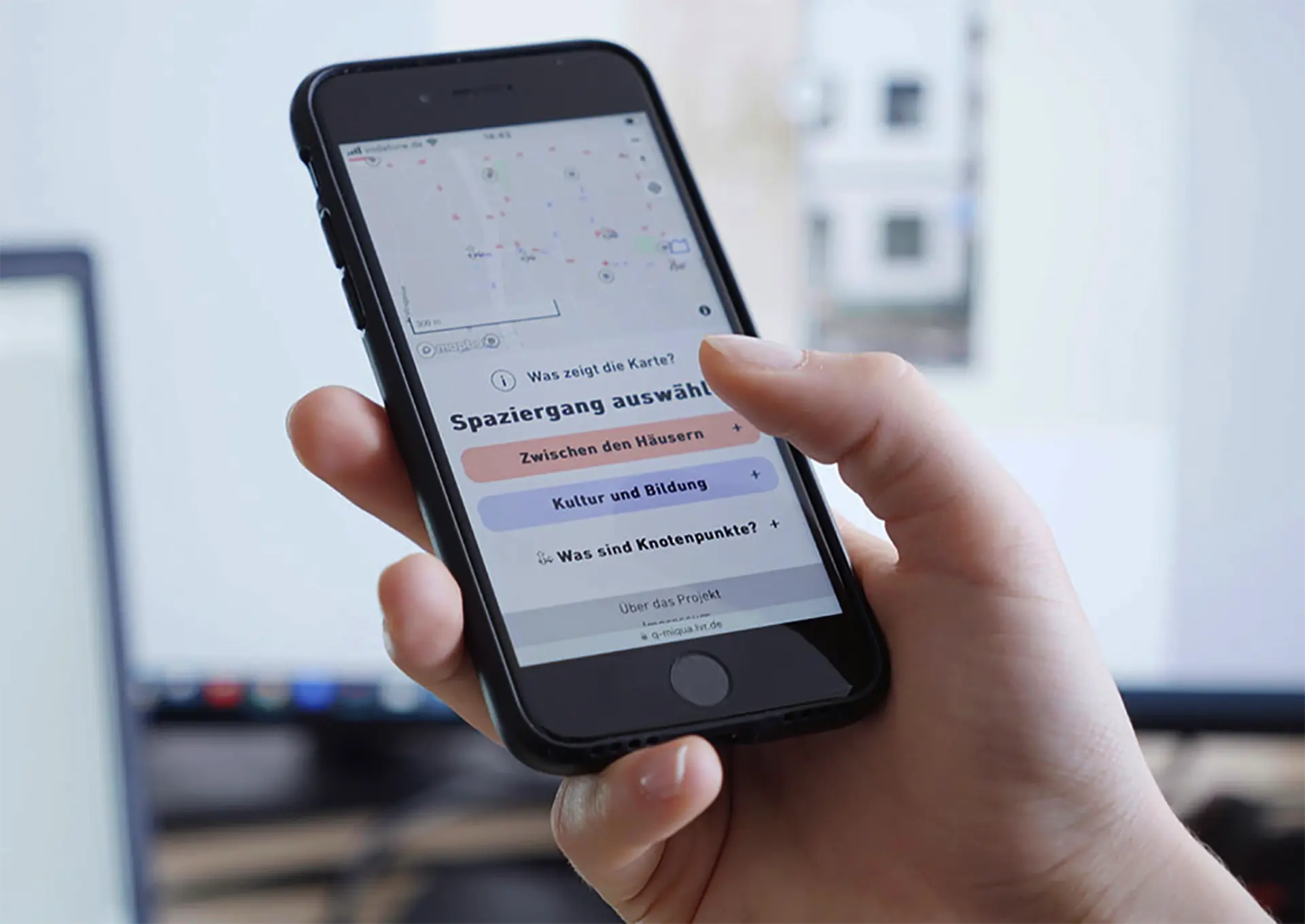 Eine Hand hält ein Smartphone mit geöffnetem Startbildschirm der Miqua-App, auf dem eine Karte und Auswahloptionen für Stadtführngen angezeigt werden.“