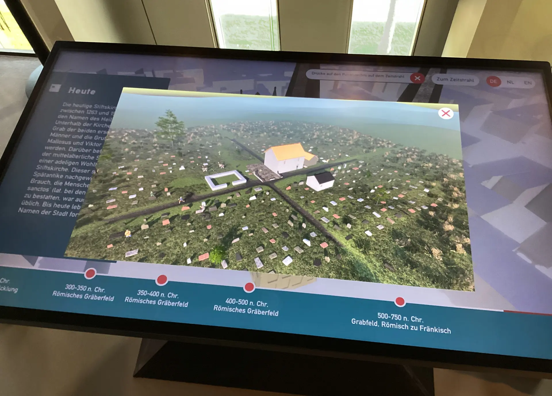Interaktiver Touchtisch mit digitaler Darstellung der Stiftskirche St. Viktor und einem Zeitstrahl zur historischen Entwicklung des umliegenden Gräberfeldes