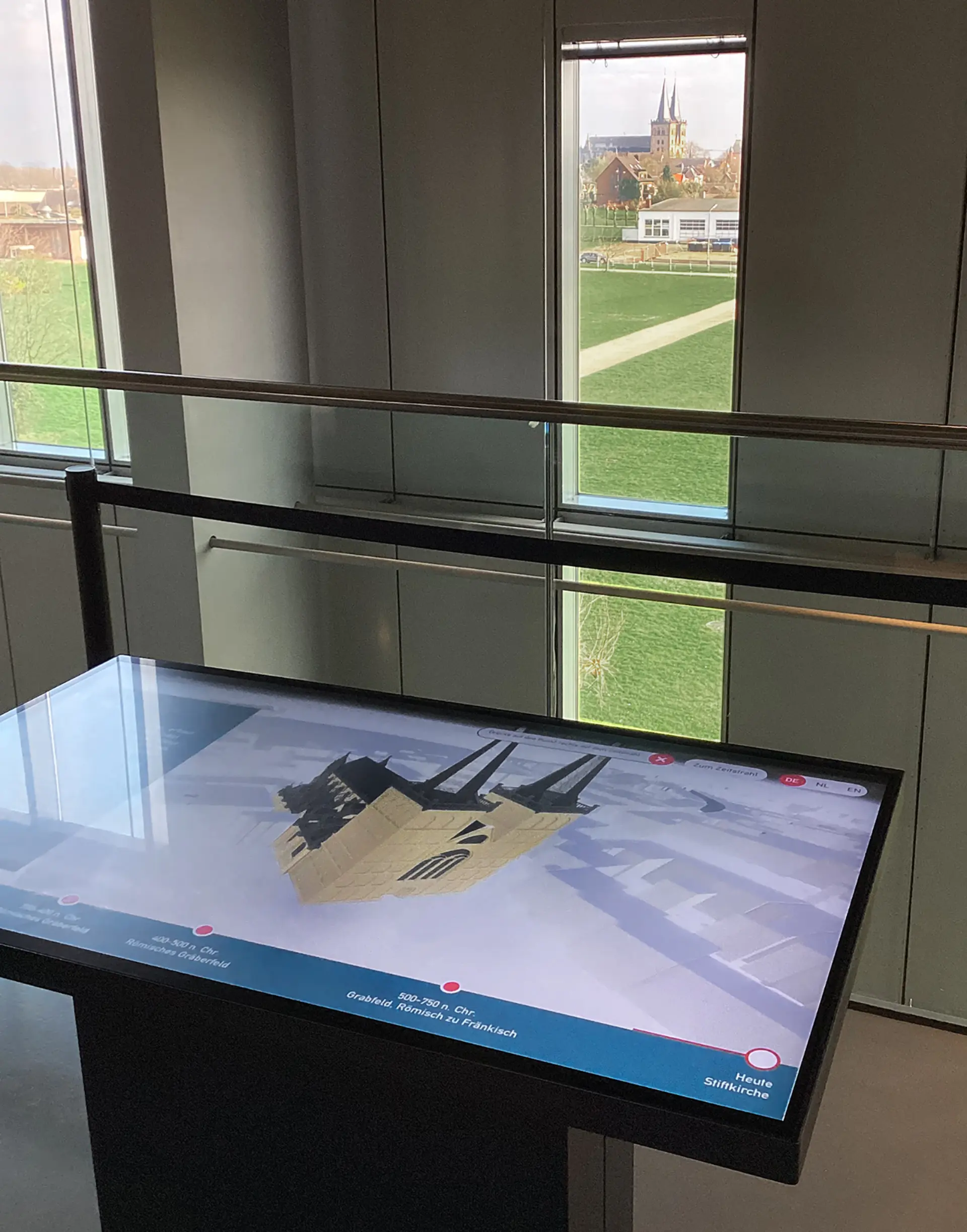 Interaktiver Touchtisch mit digitalem Modell der Stiftskirche St. Viktor am Spielende; durch die Fenster ist die reale Stiftskirche in direkter Sichtachse zu sehen