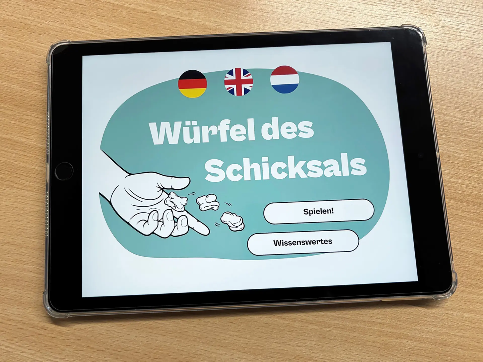 Tablet mit der Startseite der mehrsprachigen App „Würfel des Schicksals“. Zu sehen sind Sprachsymbole, eine Illustration einer würfelnden Hand sowie die Buttons „Spielen!“ und „Wissenswertes“