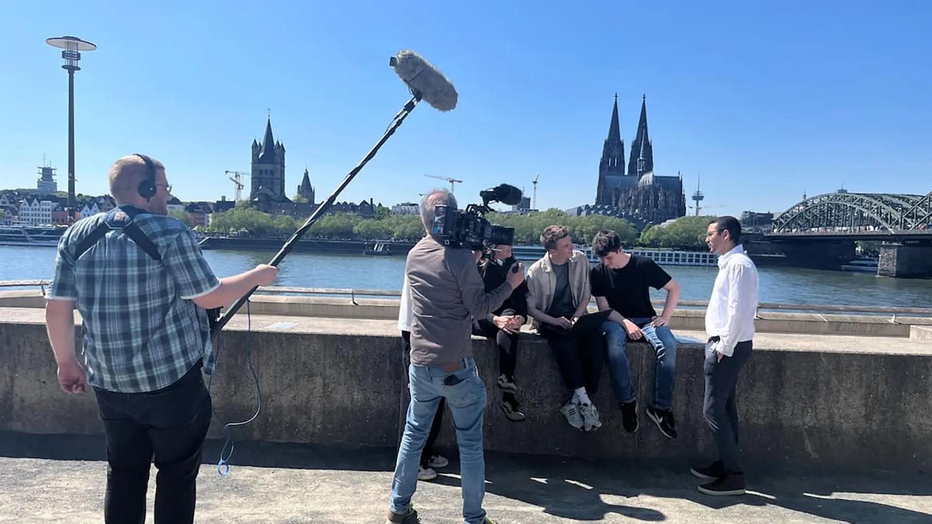 Kamerateam filmt junge Menschen, die in Köln am Rhein auf einer Mauer sitzen. Im Hintergrund sieht man den Kölner Dom.